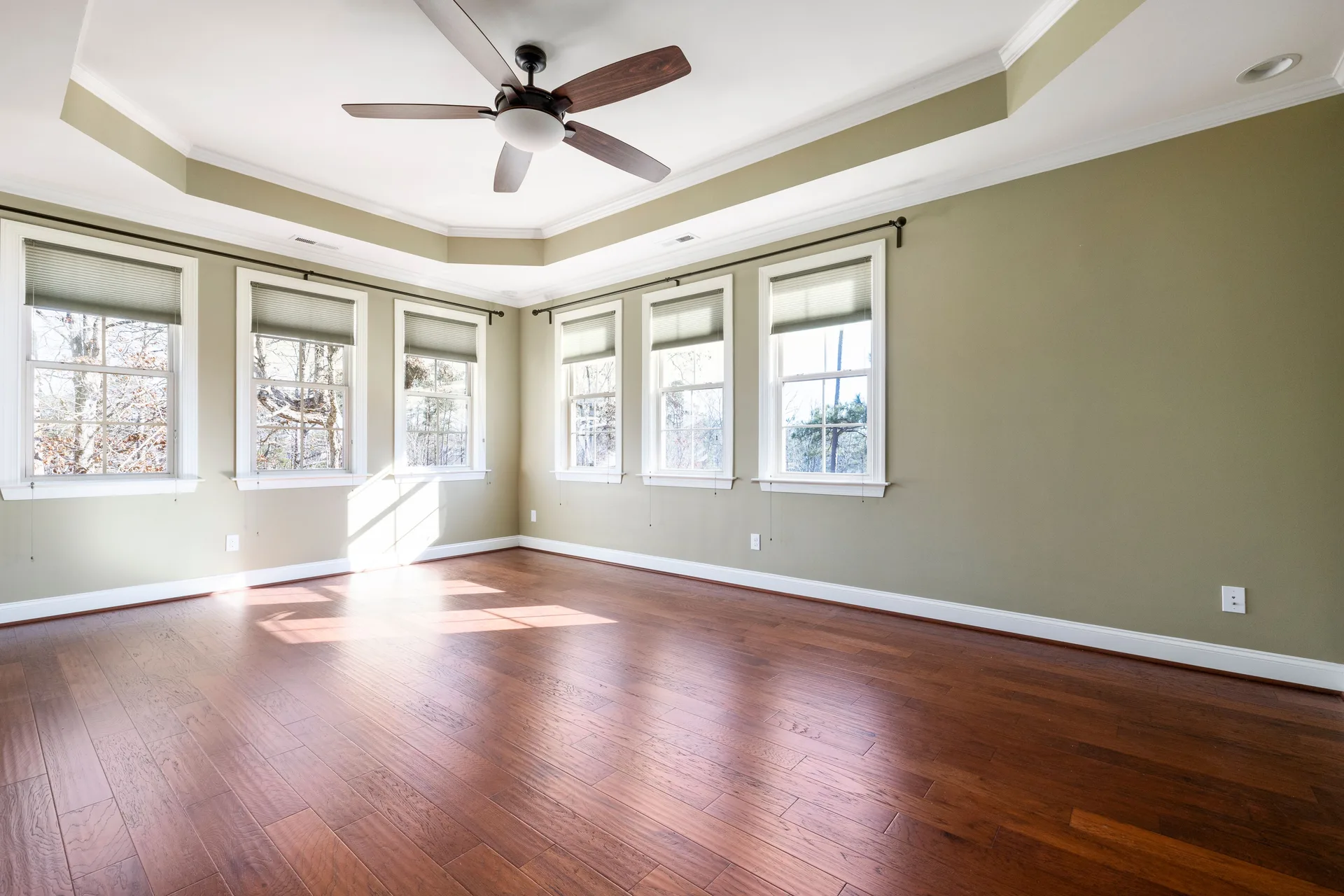 A bright empty room with a hardwood floor, large windows, and a ceiling fan