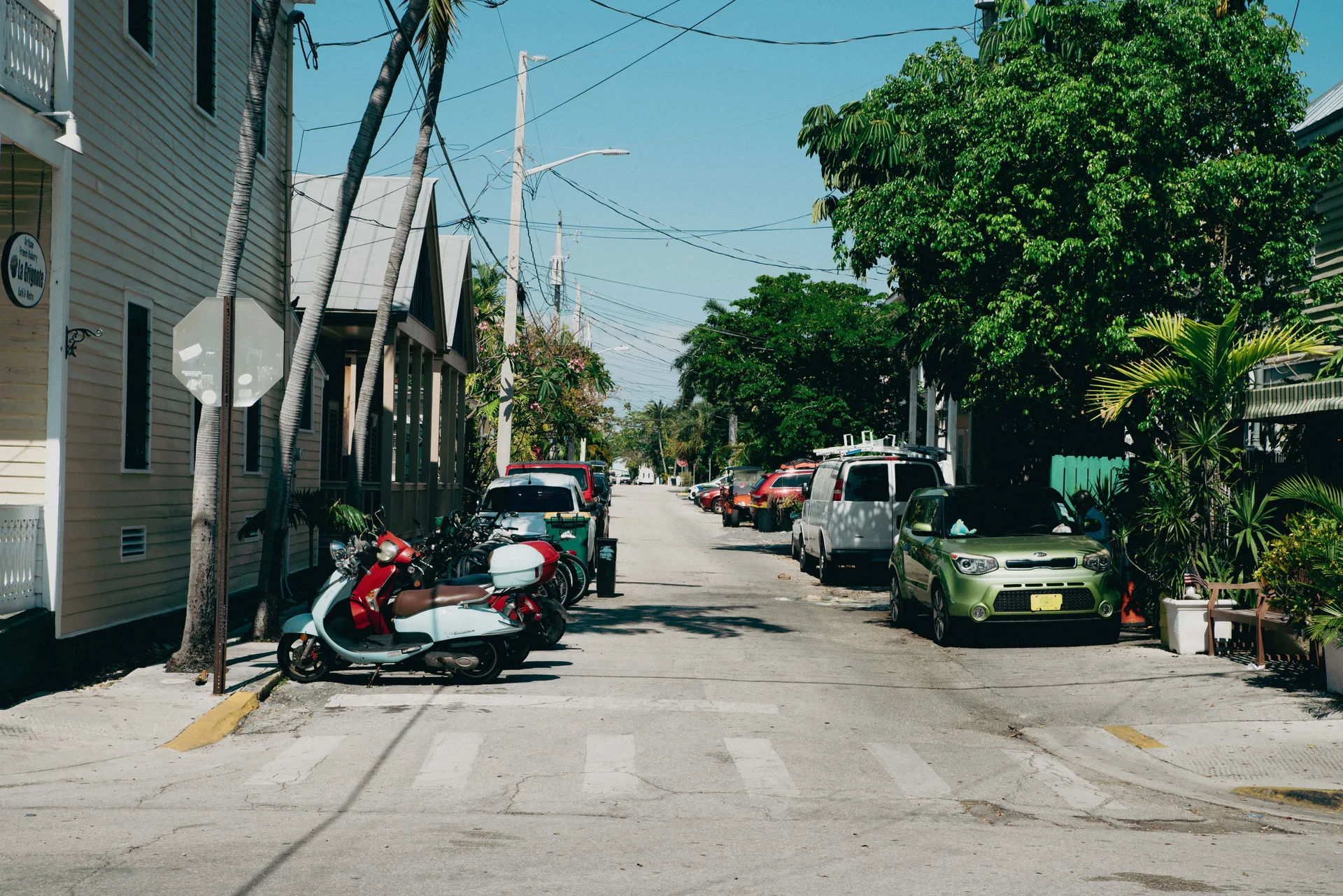 An urban street with parked cars, scooters, and greenery on a sunny day