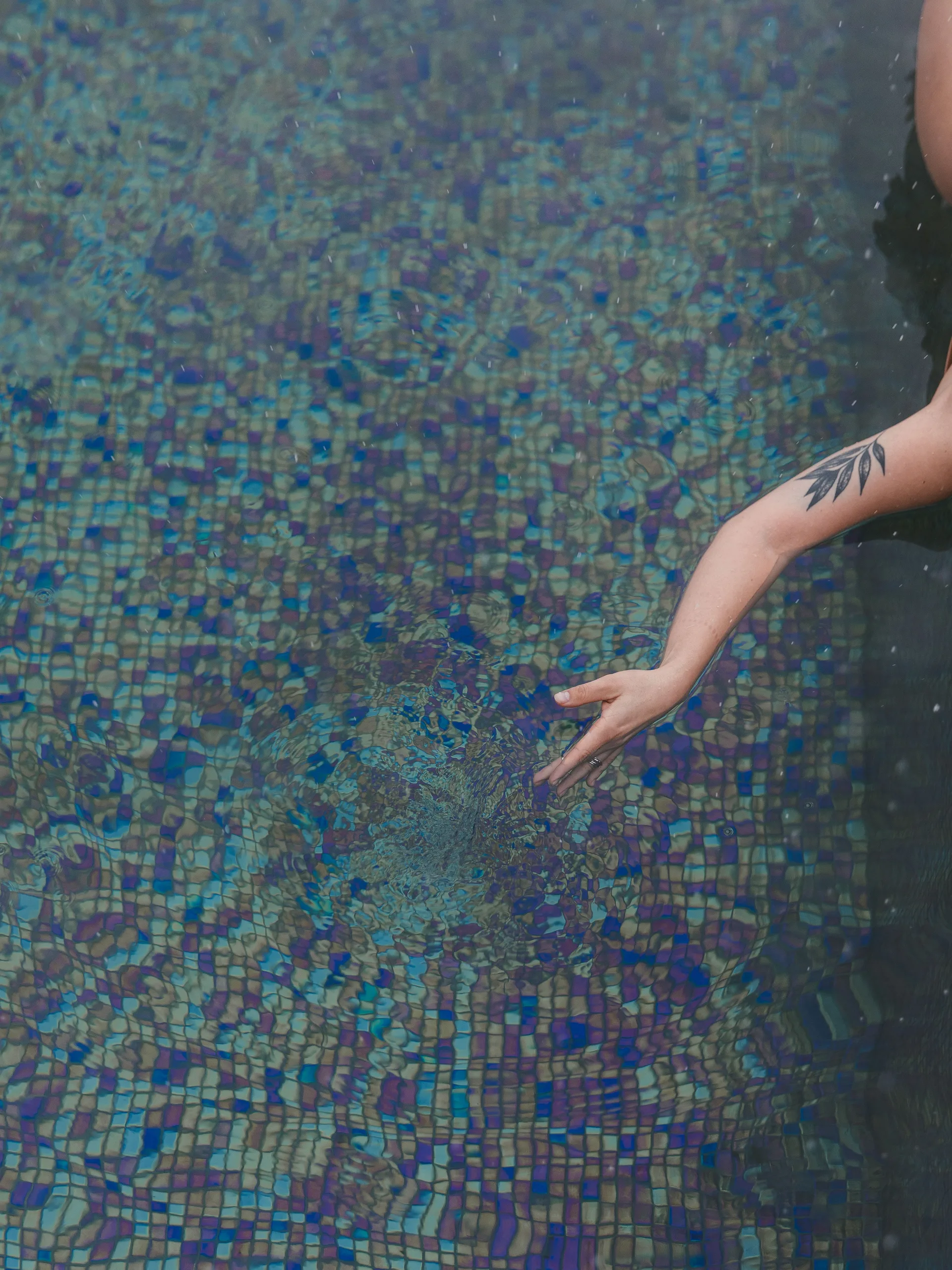 Arm reaching into clear pool water over mosaic tiles